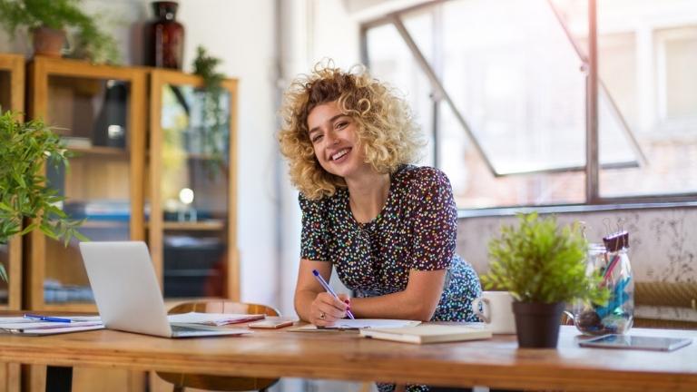A blonde female business owner works at a long desk in a light office filled with plants