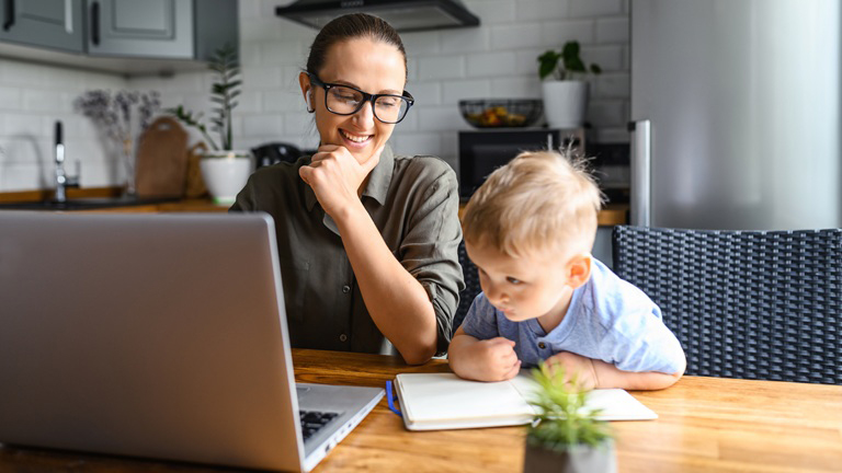 A woman works on a laptop at a kitchen table while a young child sits beside her, drawing in a notebook.