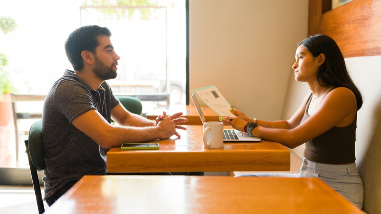 Two people sit at a wooden table in a cafe, discussing something on a laptop. A coffee mug and phone are on the table.