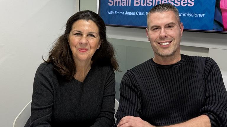 A woman and a man are seated side by side, smiling warmly. They are indoors, with a sign in the background referencing tackling late payments for small businesses.