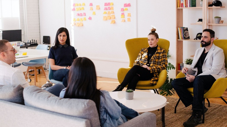 A group of five people having a discussion in a modern office with colourful sticky notes on the wall and bookshelves in the background.