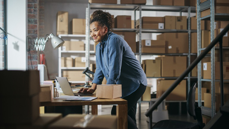 A woman in a blue shirt stands at a desk with a laptop, surrounded by shelves filled with cardboard boxes in a warehouse setting.