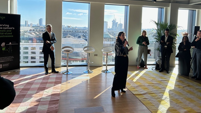 A woman speaks into a microphone in a sunlit room with a city view. People stand attentively nearby, creating a professional and engaging atmosphere.