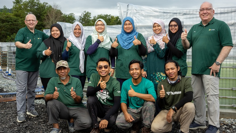 A diverse group of people smiling and giving thumbs up, standing outdoors in front of construction materials and greenery.