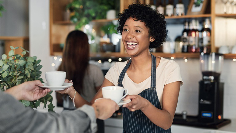 Smiling young café barista handing customer two coffee cups Barista in an apron smiling while handing a cup of coffee to a customer in a cosy café setting.