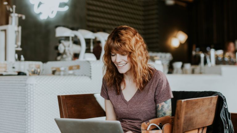 A smiling young woman with red hair sits at a table in a coffee shop working on her laptop