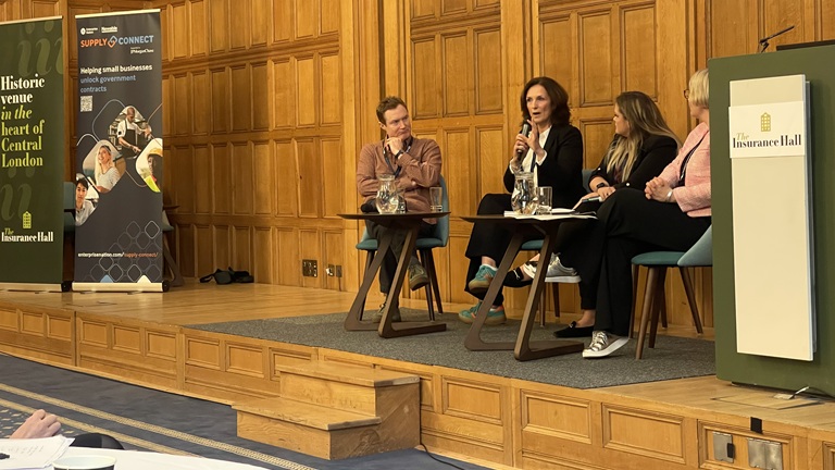 Panel discussion at a conference in a wood-panelled room, with four people seated on stage, one speaking into a microphone.