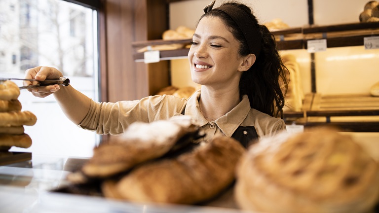 A woman working in a bakery shop, serving pastries and fresh bread with tongs