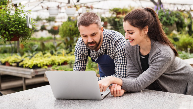 Two people in a greenhouse, looking at a laptop. The woman points at the screen while the man leans in. Plants are visible in the background.
