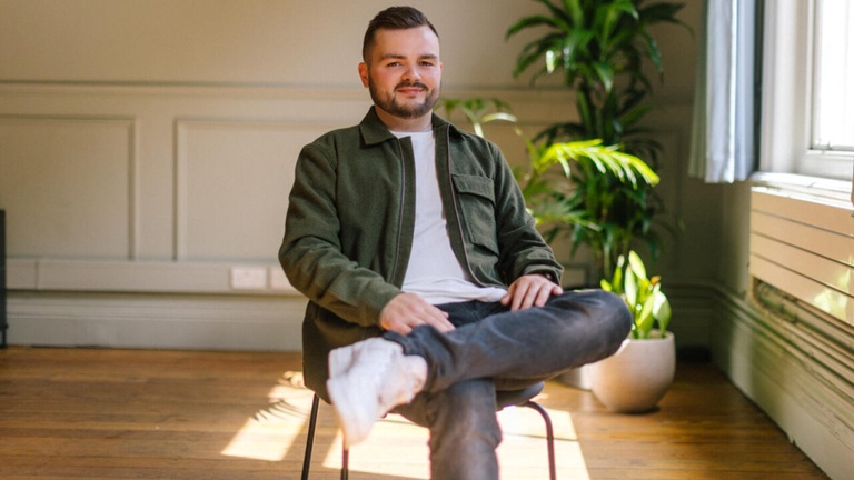 A man sits casually on a chair in a sunlit room with wooden flooring, crossed legs, wearing a green jacket. A large potted plant is in the background.