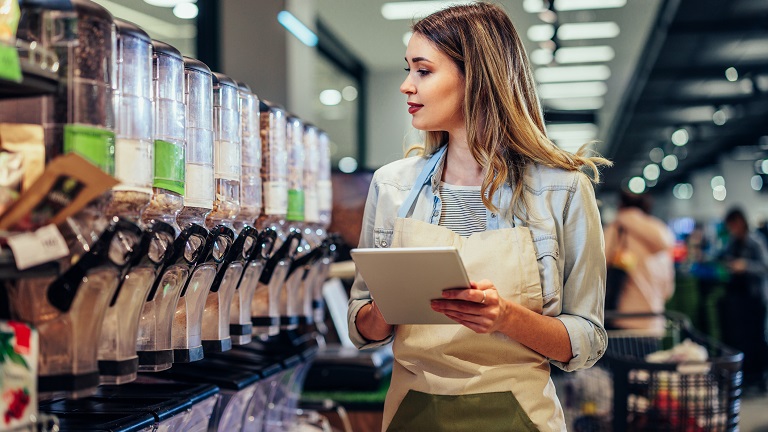 Young female shop worker checking containers of dried foods against notes on a tablet