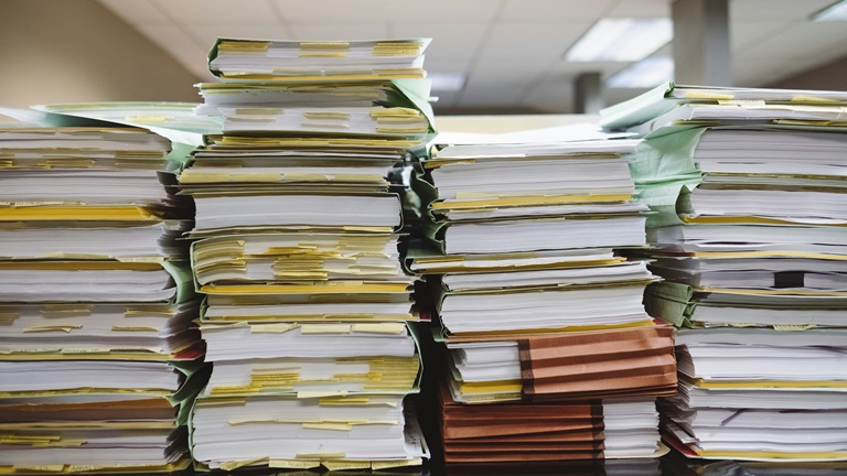 Stacks of paper folders and documents piled high on a desk, with yellow sticky notes attached, under fluorescent lighting.