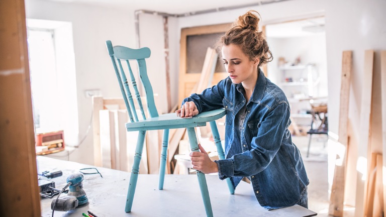 A young white female carpenter sands a turquoise wooden chair atop a workbench