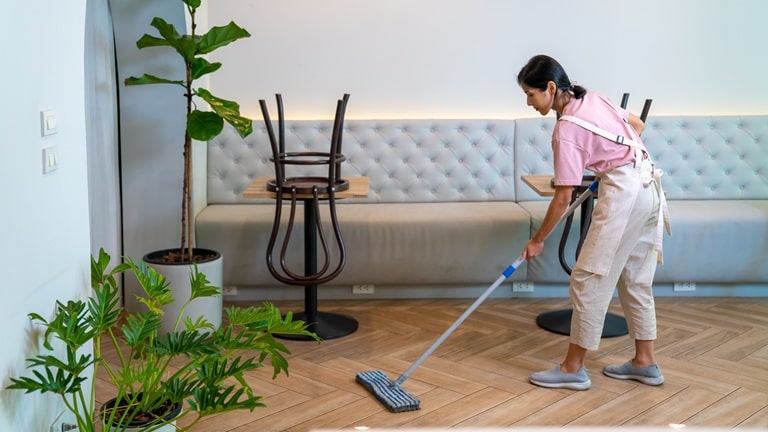 A person mopping a wooden floor in a cafe, with chairs stacked on tables and green plants nearby.