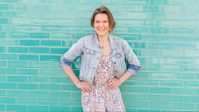 Smiling woman in a floral dress and light denim jacket standing with hands on hips against a teal tiled wall.