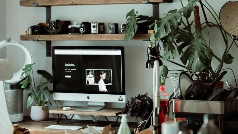 An Apple monitor and computer set up on a modern wooden desk unit, surrounded by plants