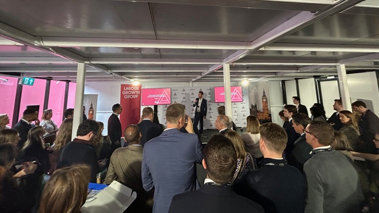A speaker addresses a crowd at an indoor event with a "Labour Growth Group" banner and a backdrop featuring Big Ben.