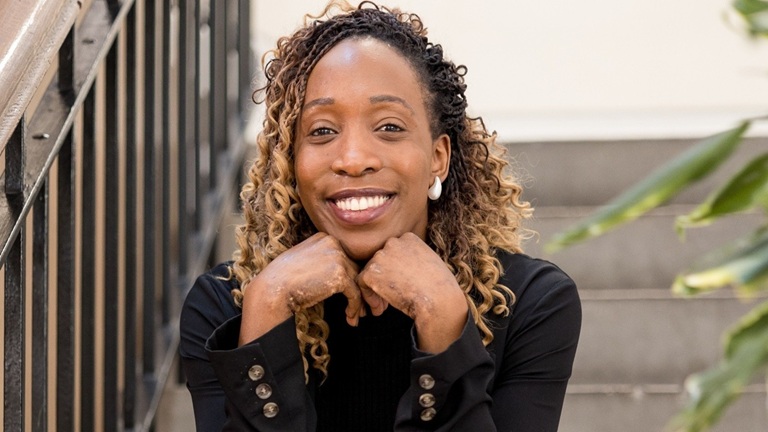 A smiling Black woman with curly hair, wearing a black top, leaning on a railing beside stairs, with a plant nearby.