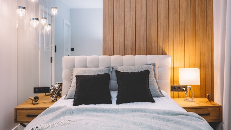 A cosy bedroom with a white tufted headboard, wooden panel wall, two black pillows, a bedside lamp and decorative items on wooden nightstands.