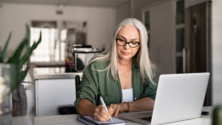 A middle-aged fashionable woman sitting at her desk at home, writing on a notepad beside her laptop