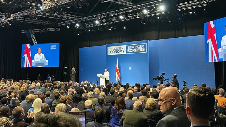 A speaker addresses a large audience at a conference, with banners reading "Stronger Economy" and "Stronger Borders" on the stage backdrop.