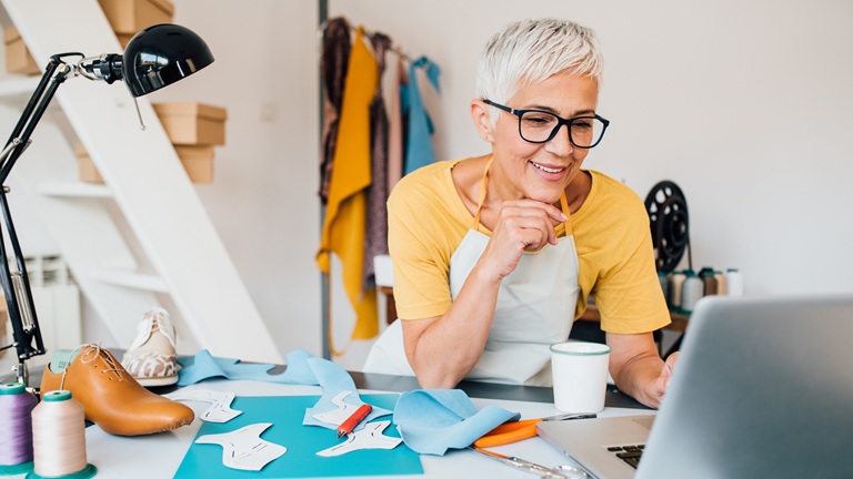 Woman with short grey hair and glasses works on a laptop in a craft room, surrounded by sewing materials and a shoe.