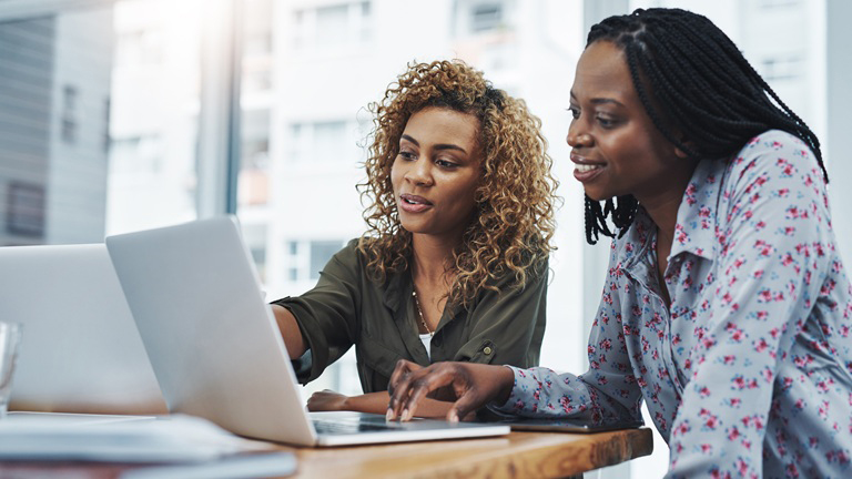 A Black female mentor sitting beside a Black female small business owner as they discuss things on the laptop between them