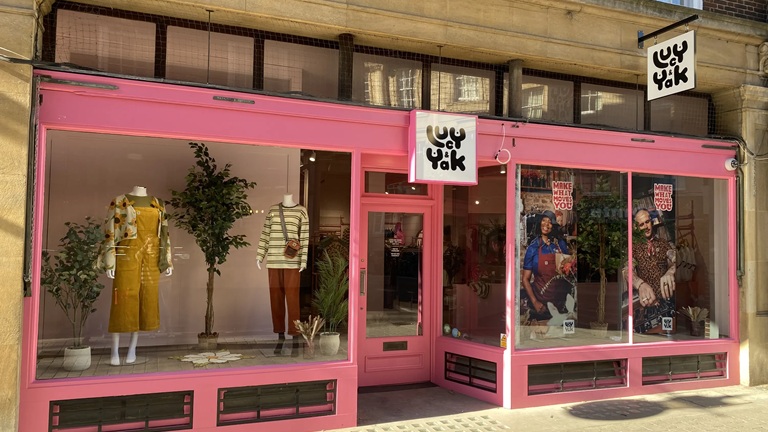 A bright pink storefront with mannequins in colourful outfits and plants inside. Signs display the store's name and promotional posters.