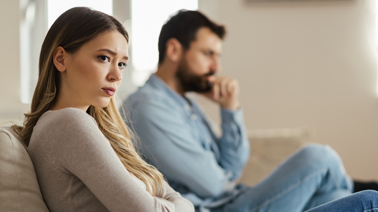 A young woman sits on a couch, looking upset, with a man in the background appearing contemplative. The scene conveys tension and conflict in a relationship.