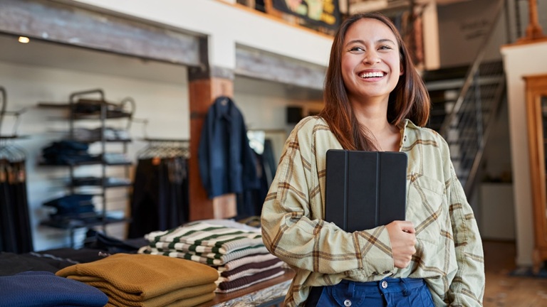 Female clothes shop owner smiling and holding tablet Smiling woman holding a tablet in a clothing store, standing near folded sweaters on a wooden table.