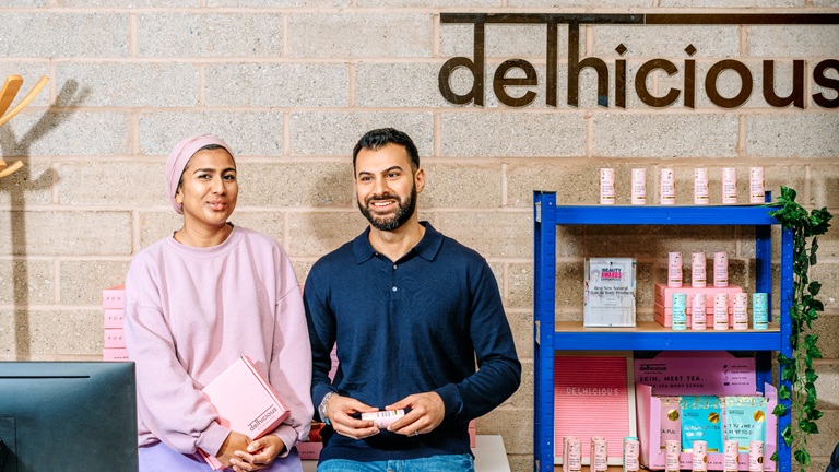 Two Asian people smiling, standing in a shop with "delhicious" sign, holding products. Shelves display pink and white boxes. Brick wall background.