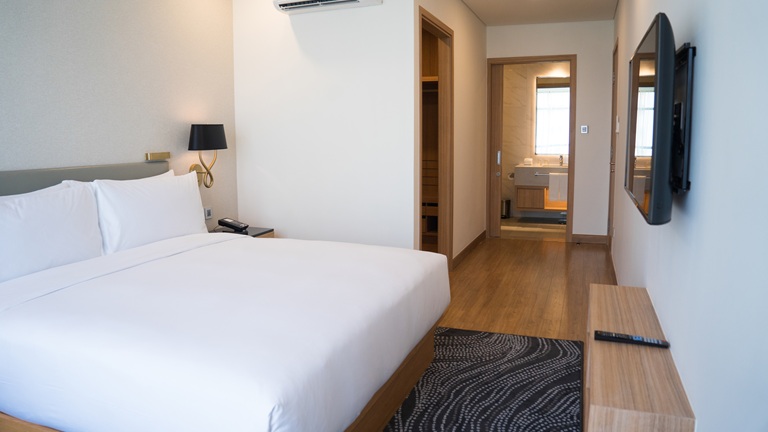 Modern hotel room with a large bed, wall-mounted TV, wooden flooring, and an open door leading to a bathroom with a visible sink.