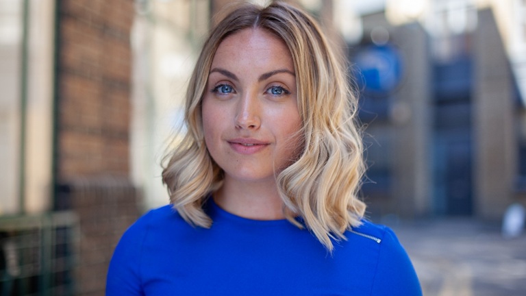 A smiling white woman with wavy blonde hair in a blue shirt stands outdoors. The background is blurred, showing brick walls and glass.
