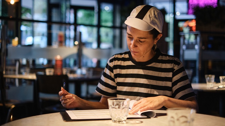 A female restaurant owner in white cap and striped T-shirt sits at a table doing her accounts on paper