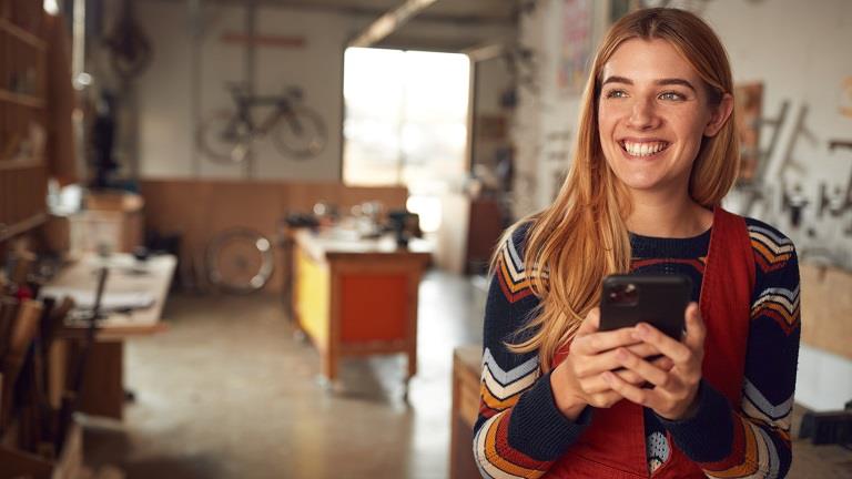 A young white female business owner in her workshop, holding a smartphone and smiling