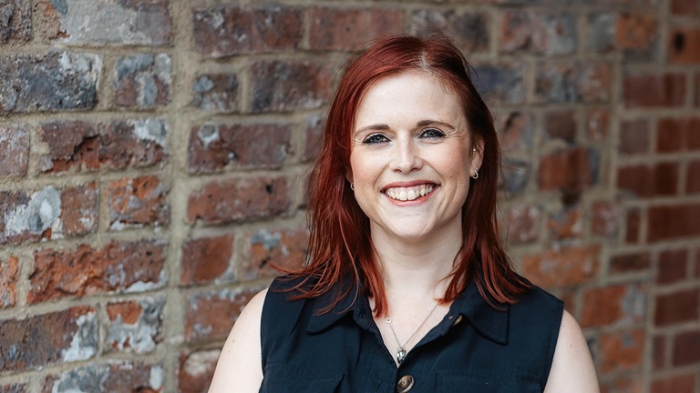 Smiling red‑haired woman in a sleeveless dark blouse standing against a weathered brick wall.