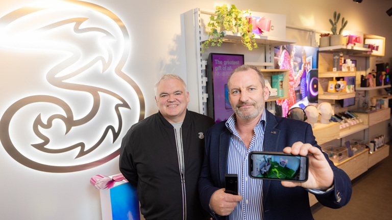 Two men in a tech store, one holding a smartphone displaying a colourful screen. A glowing "3" logo is on the wall behind them.