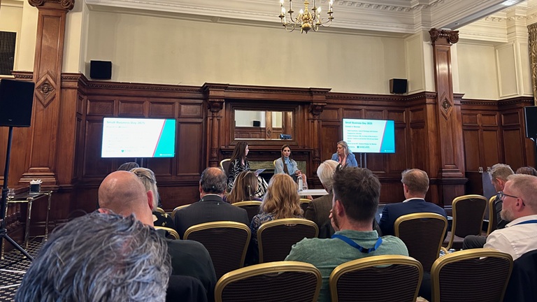 Audience attending a panel discussion in a wood-panelled room with two speakers and presentation screens displaying slides.