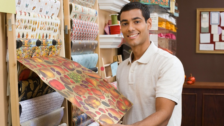 A young Asian male owner in a white polo shirt smiles and holds out a sheet of wrapping paper from a rack on the wall