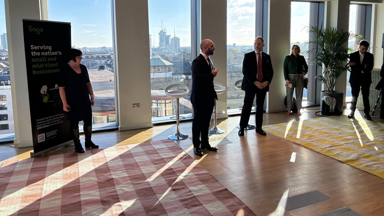 A group of people stand in a sunlit room with large windows showing a cityscape. Two men in suits are speaking near a banner about supporting businesses, with others listening.