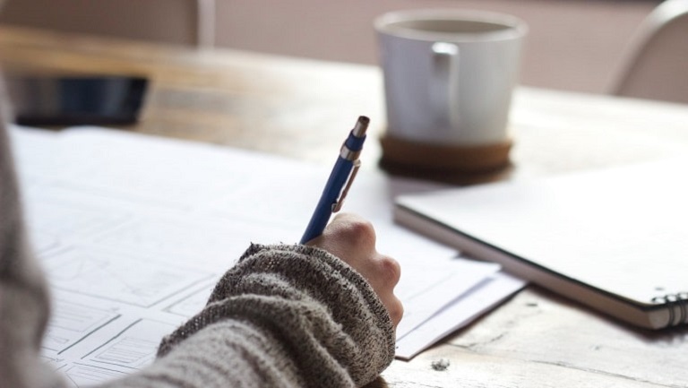 A woman's hand holding a pen and filling in tax paperwork and business records laid out on a table