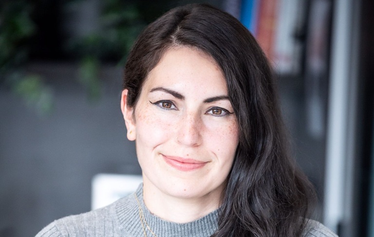 A woman with long dark hair and winged eyeliner smiles softly. She wears a grey sweater against a blurred background of books and greenery.