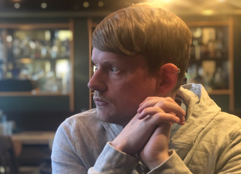 A young white man with light brown hair is gazing thoughtfully to the side, seated in a cosy, softly lit bar. He has clasped hands and wears a light-coloured hoodie.