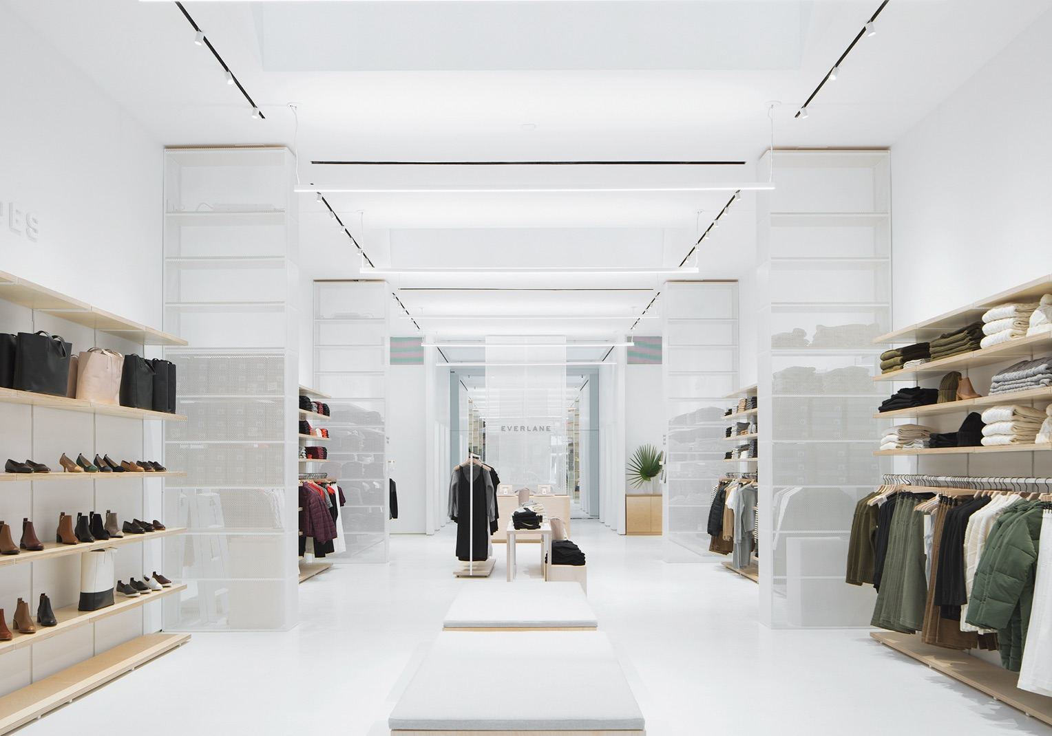 Inside view of Everlane's store. The room is mainly white with wooden System 1224 shelves along the walls displaying a variety of clothes.