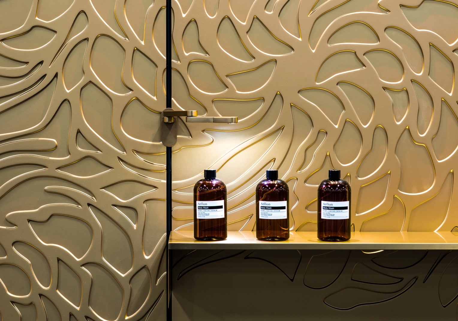 Close up shot of a golden floral patterned and metallic wall. A golden shelf along it is holding up three brown glass bottles.