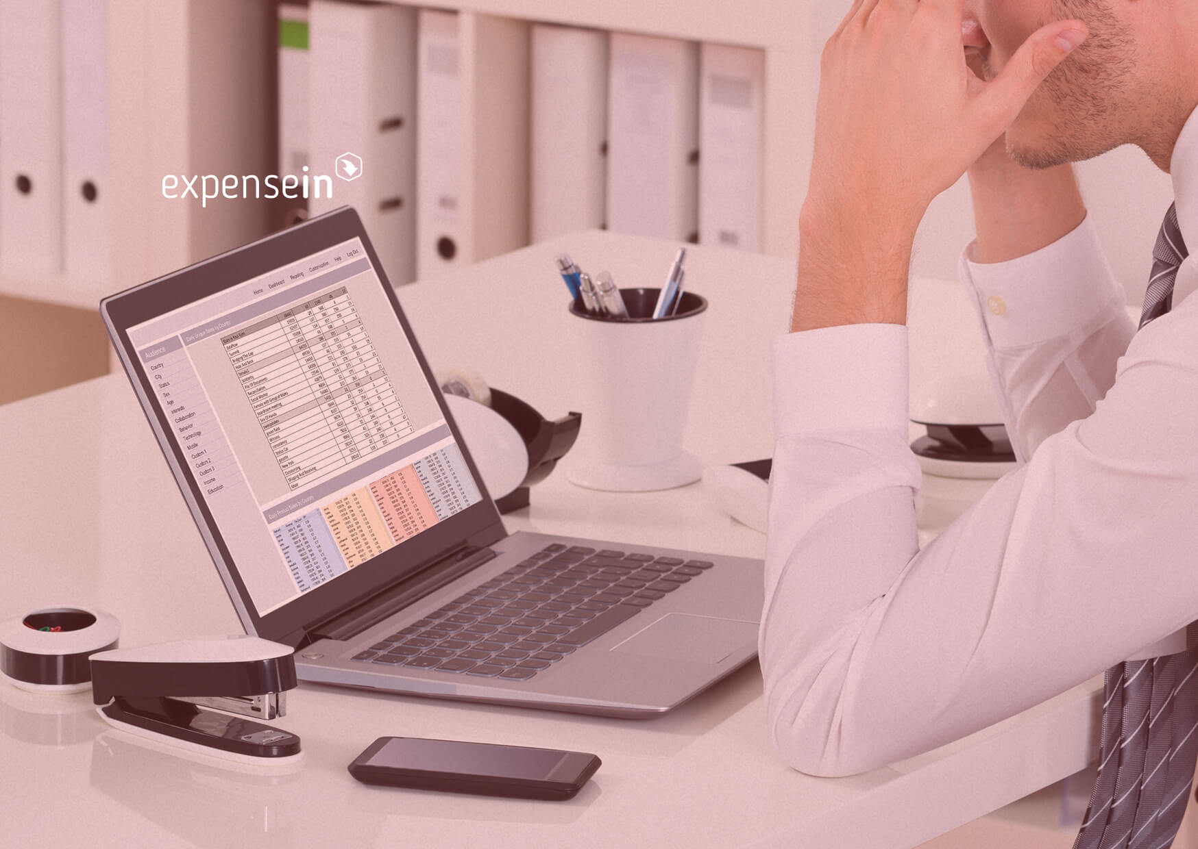 Person at a desk using a laptop displaying a spreadsheet. Nearby are office supplies like a stapler and smartphone.