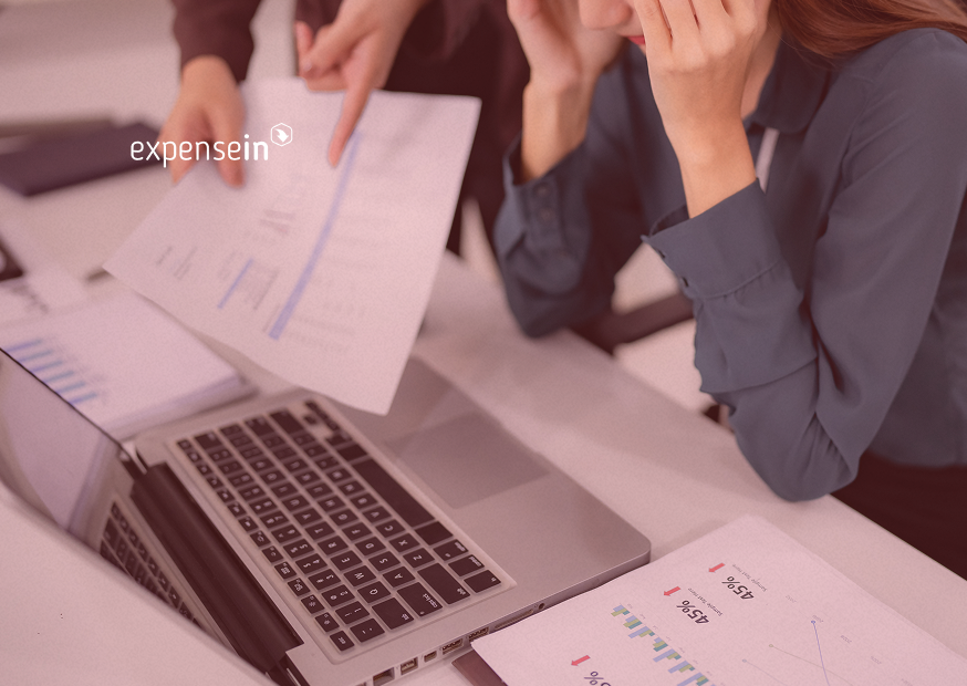 Two people reviewing documents over a laptop with charts and graphs visible. One person holds a paper, and the other rests their head in their hands.