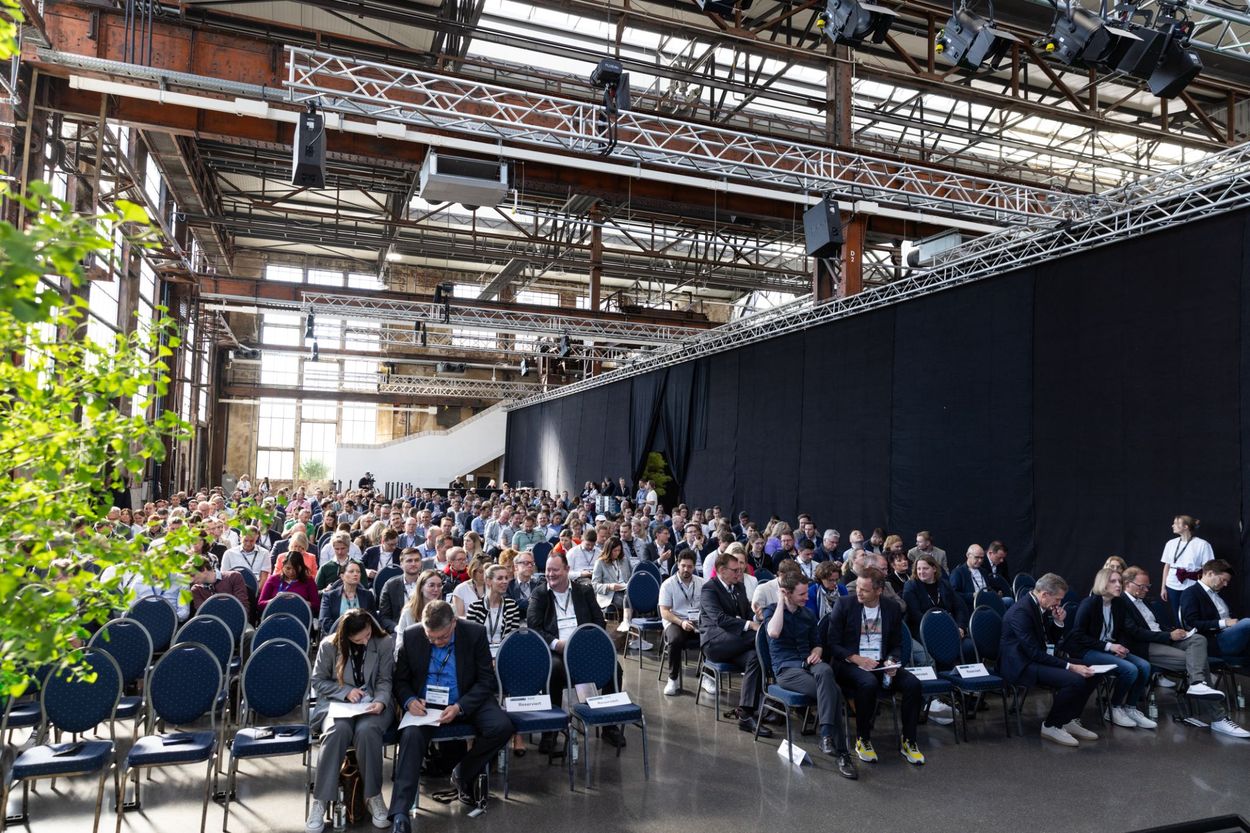 A large audience seated in rows at a conference in an industrial-style venue with high ceilings and large windows.