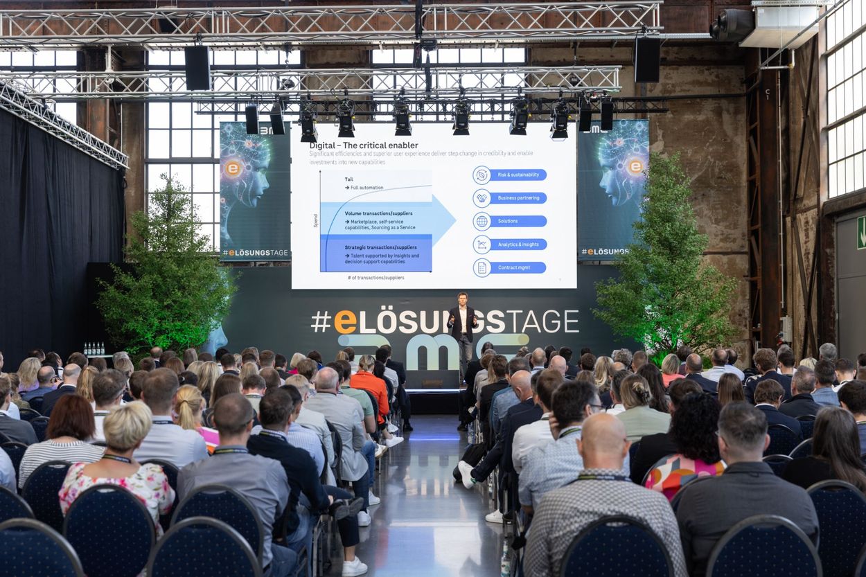 A speaker presents to a seated audience on digital transformation at a conference, with a large screen displaying a diagram in the background.