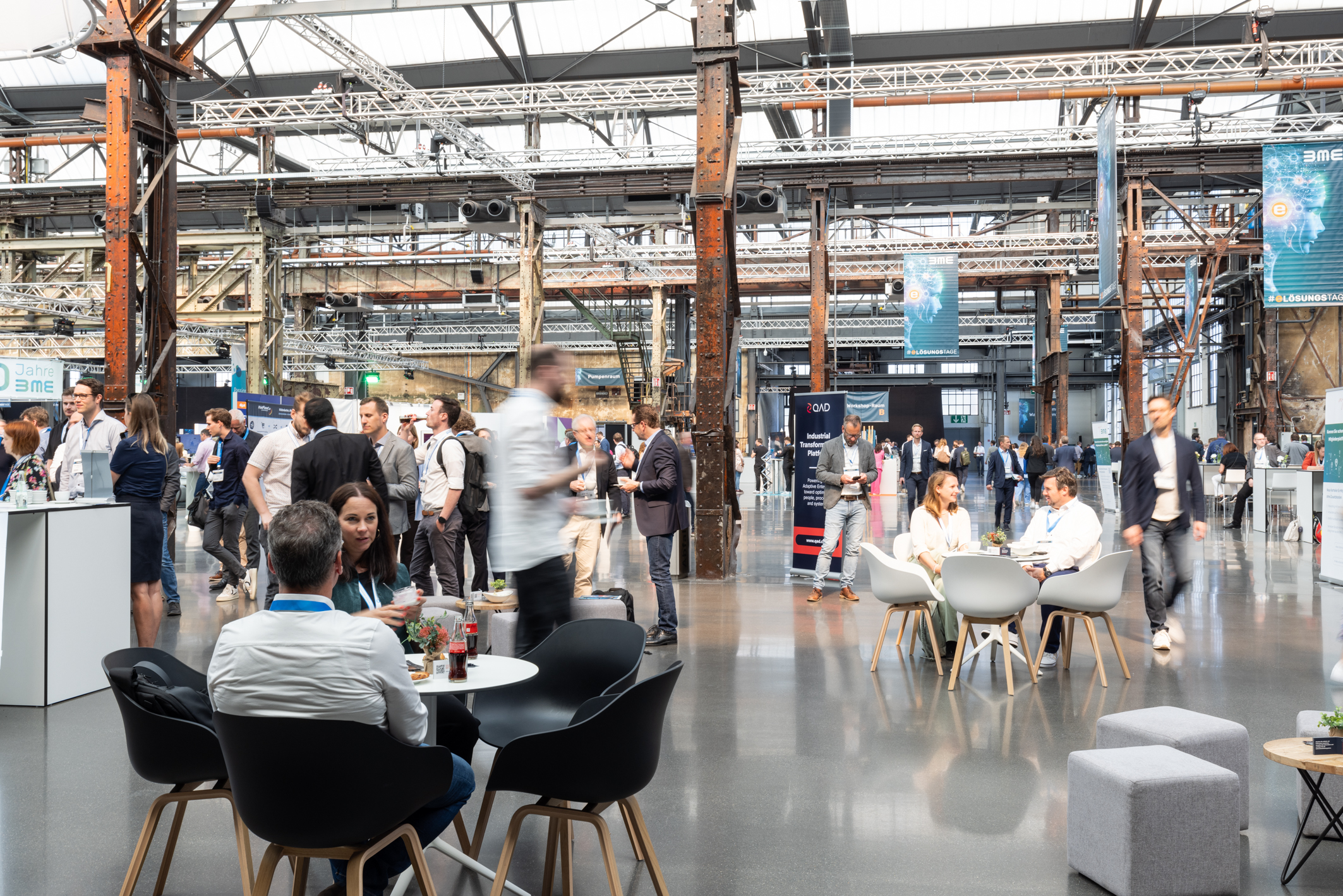 People engaging at a modern industrial-style conference venue, with attendees sitting and walking amidst exhibits and networking areas.